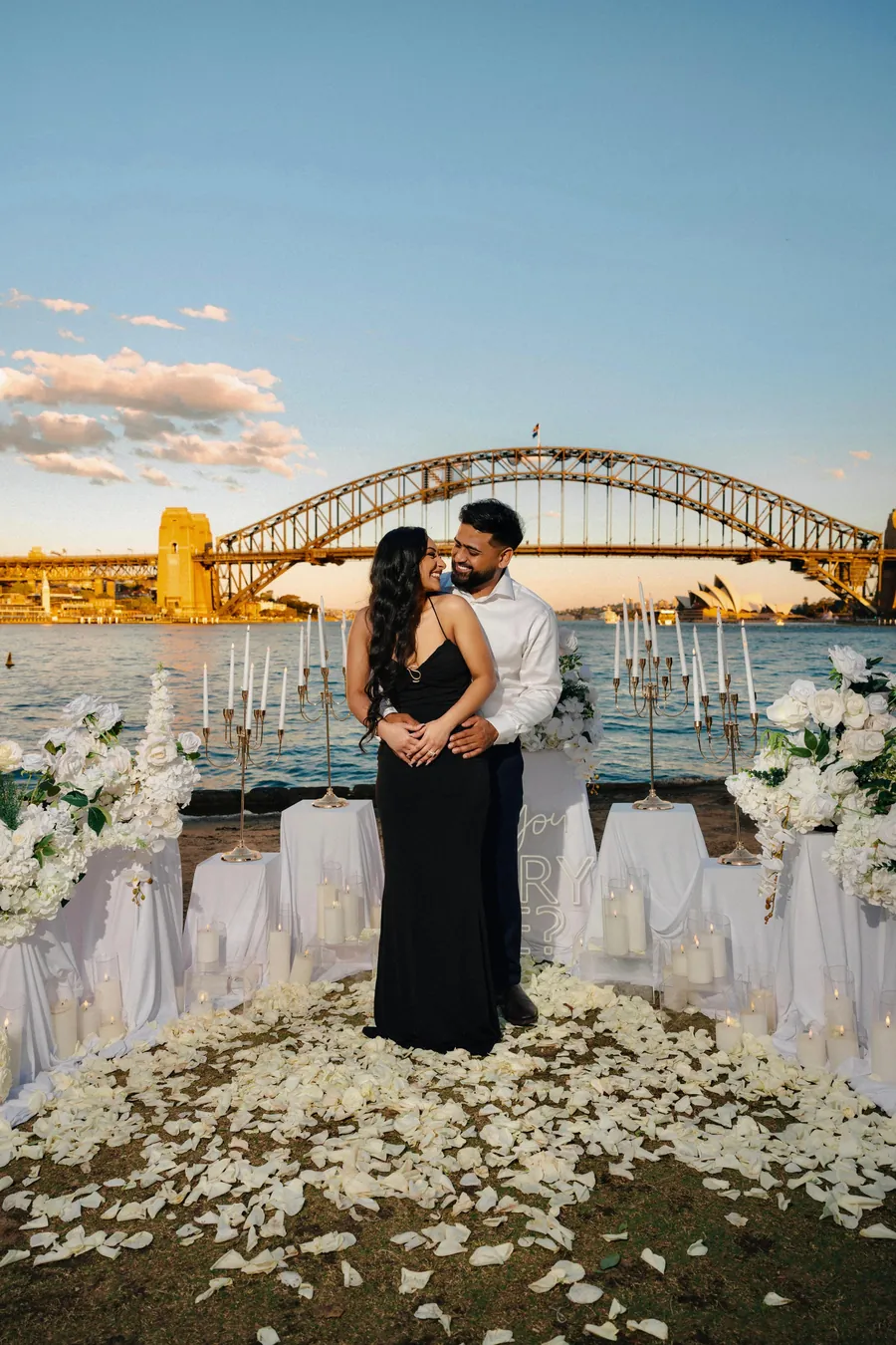 Jaz and Rianna embracing among white flowers by the Harbour Bridge